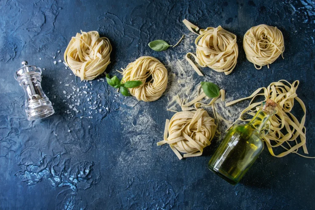 Fresh handmade pasta nests with basil, olive oil, and sea salt on a dark blue textured surface — authentic handmade pasta in Singapore.
