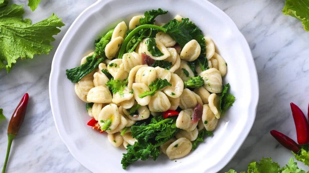 Orecchiette con Cime di Rapa on a white plate placed on a marble countertop with red peppers and lettuce leaves on the side